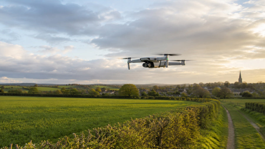 A drone hovering low over open fields beside a hedgerow, a village behind